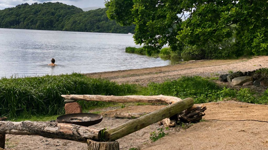 Loch Lomond Waterfront's private beach with a person wild swimming and the seating area and fire pit in the foreground