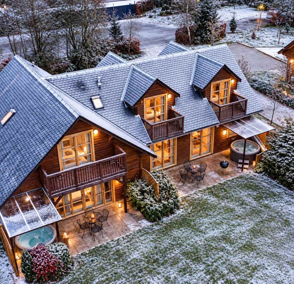 An overhead dron shot of Loch Lomond Waterfronts luxury lodges and hot tubs in winter with frost on the ground and cosy lights on inside.